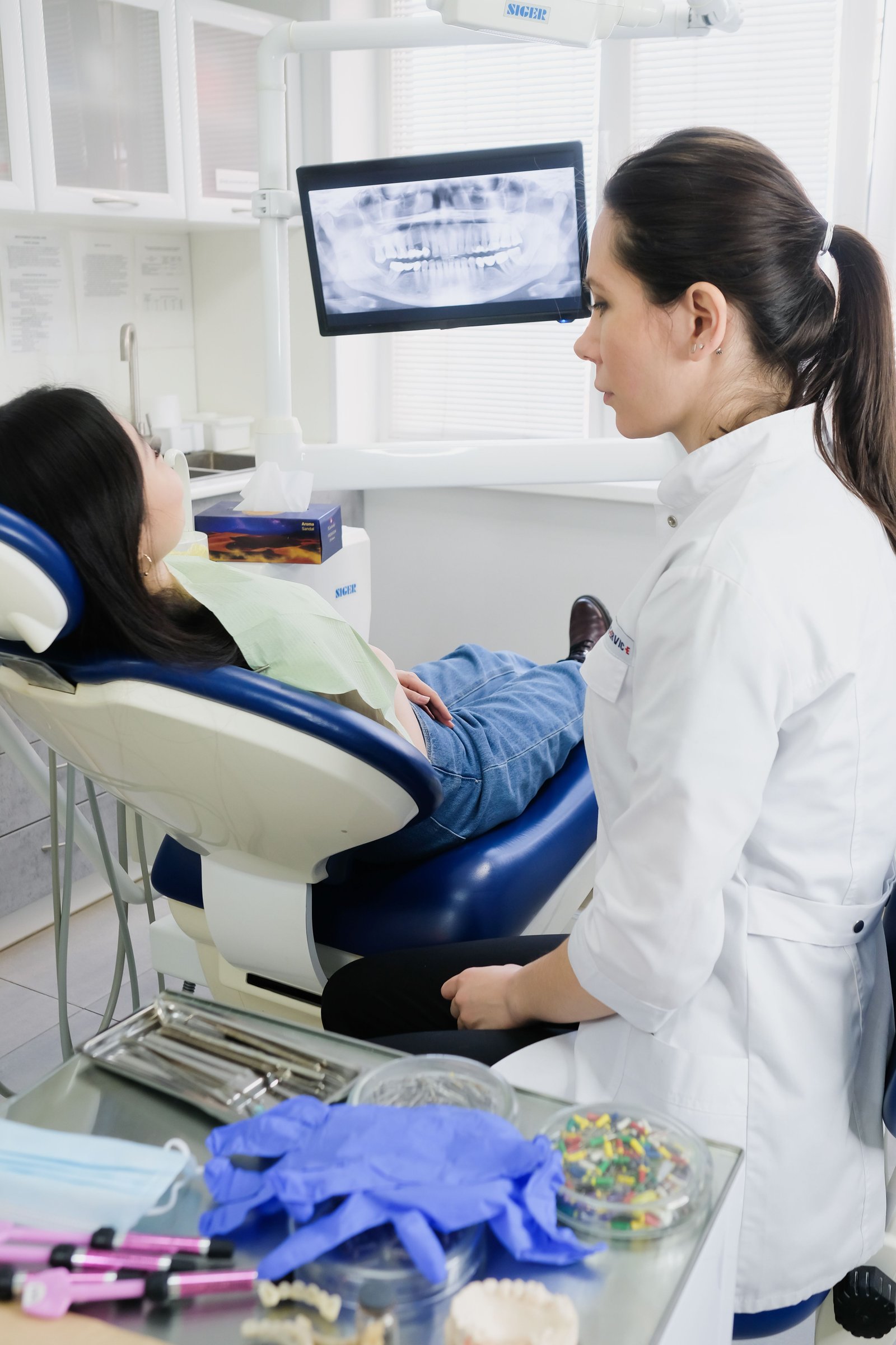 A dentist standing beside a patient in the chair, with an X-ray monitor behind