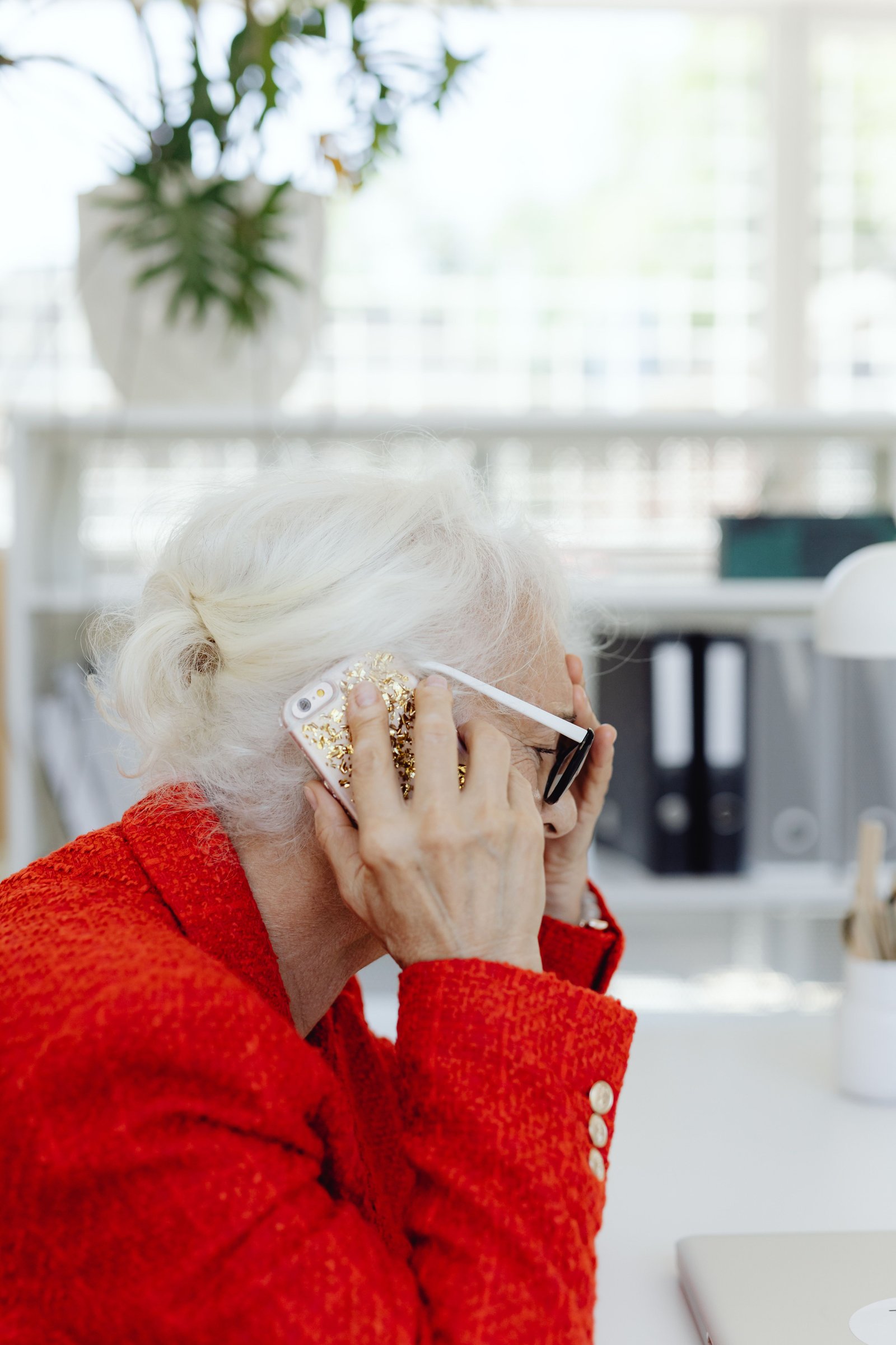 An older woman in a red jacket on the phone at her desk